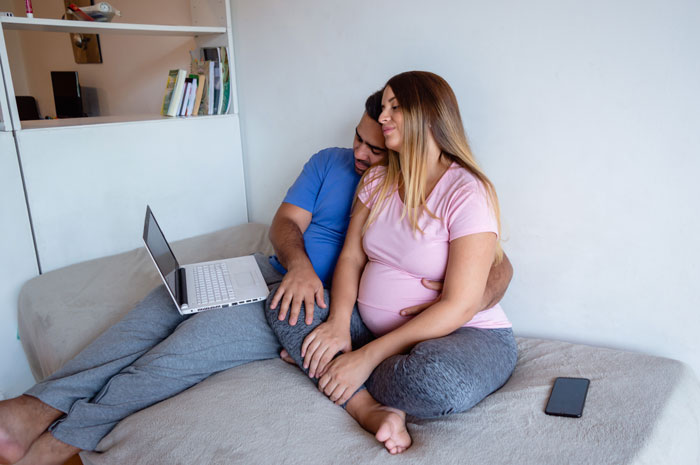 Couple sitting on a bed with laptop, man hugging pregnant woman, illustrating in-laws' misconception about weight and divorce. Couple sitting on a bed with laptop, man hugging pregnant woman, illustrating in-laws' misconception about weight and divorce.