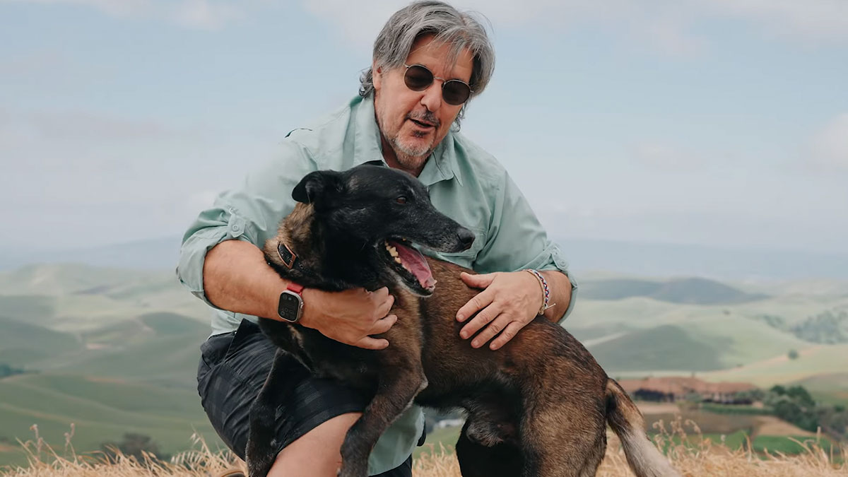 Man wearing sunglasses hugging loyal dog outdoors with green hills in the background, showcasing herou2019s smile and bond.