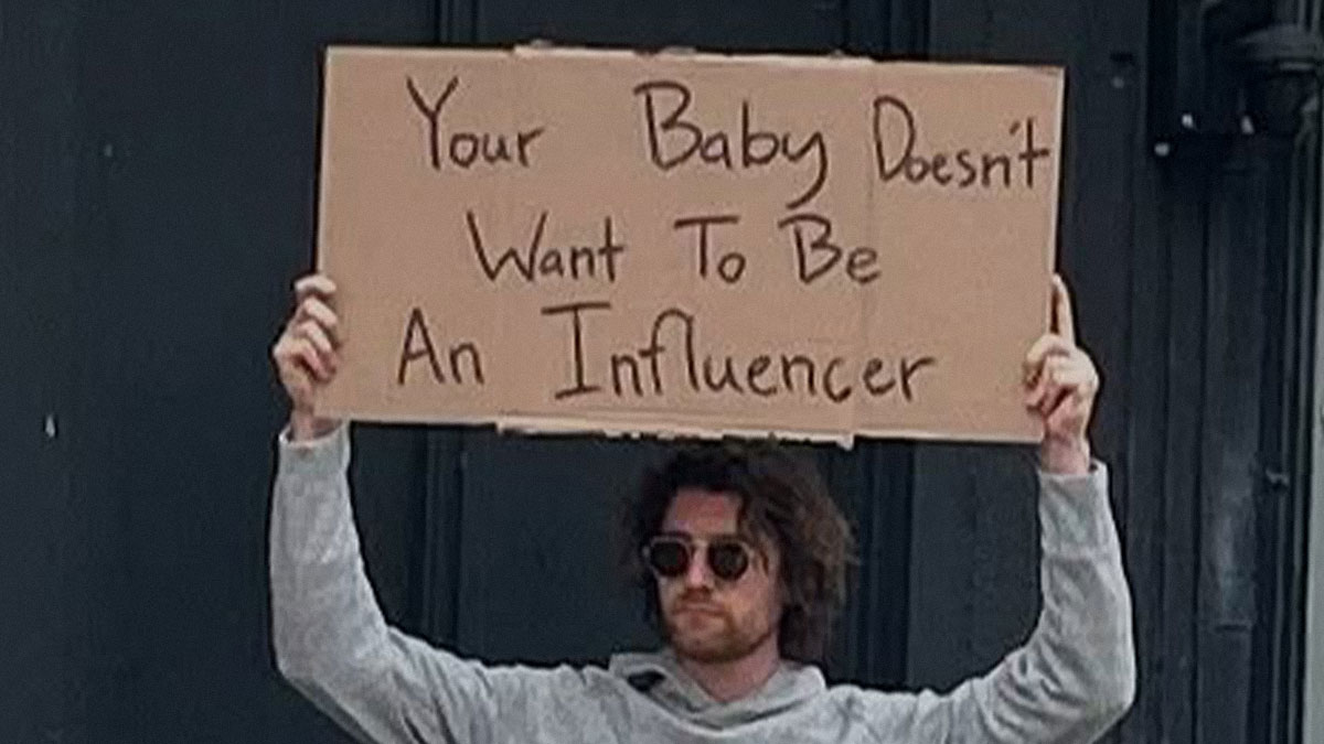 Man holding cardboard sign saying your baby doesnt want to be an influencer in a casual outdoor setting dude with the sign
