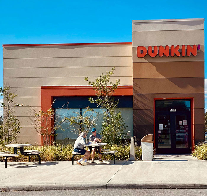 Dunkin store exterior with two people sitting outside at a table amid controversy over genetics and woke outrage. Dunkin store exterior with two people sitting outside at a table amid controversy over genetics and woke outrage.