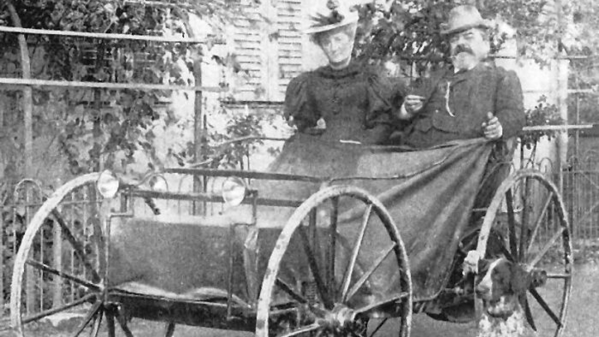 Vintage photo of two people seated in one of the first cars showing how wild early car designs were 100 years ago.