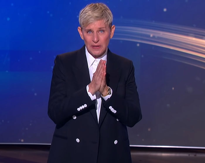 Ellen DeGeneres in a black suit, with hands clasped, addressing audience amid bullying controversy claims on stage.