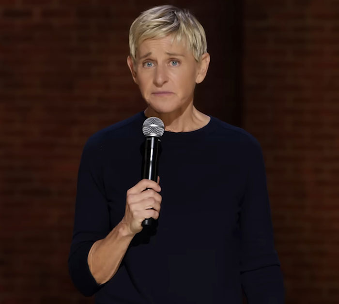 Ellen DeGeneres holding a microphone on stage, looking serious amid the bullying controversy and new claims.