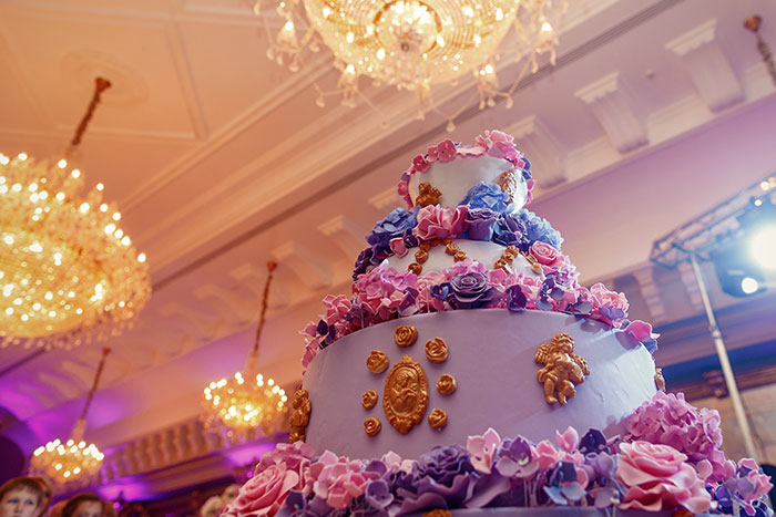 Multi-tiered decorated cake with purple and pink flowers under ornate chandeliers at a formal event setting.