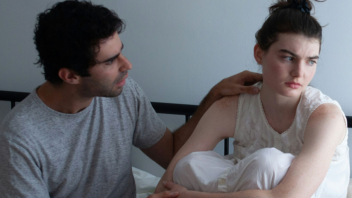 Couple having a tense conversation on a bed, woman upset while man tries to comfort her amid relationship conflict.