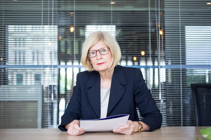Older woman in business attire sitting at desk holding papers, representing coworker retirement party and forced quit scenario Older woman in business attire sitting at desk holding papers, representing coworker retirement party and forced quit scenario
