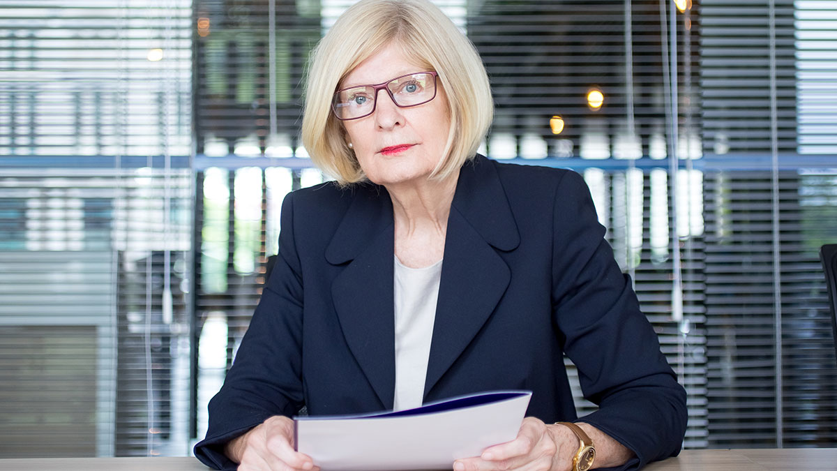 Older woman in office setting, wearing glasses and blazer, appearing serious with documents in hand, coworker retirement party concept
