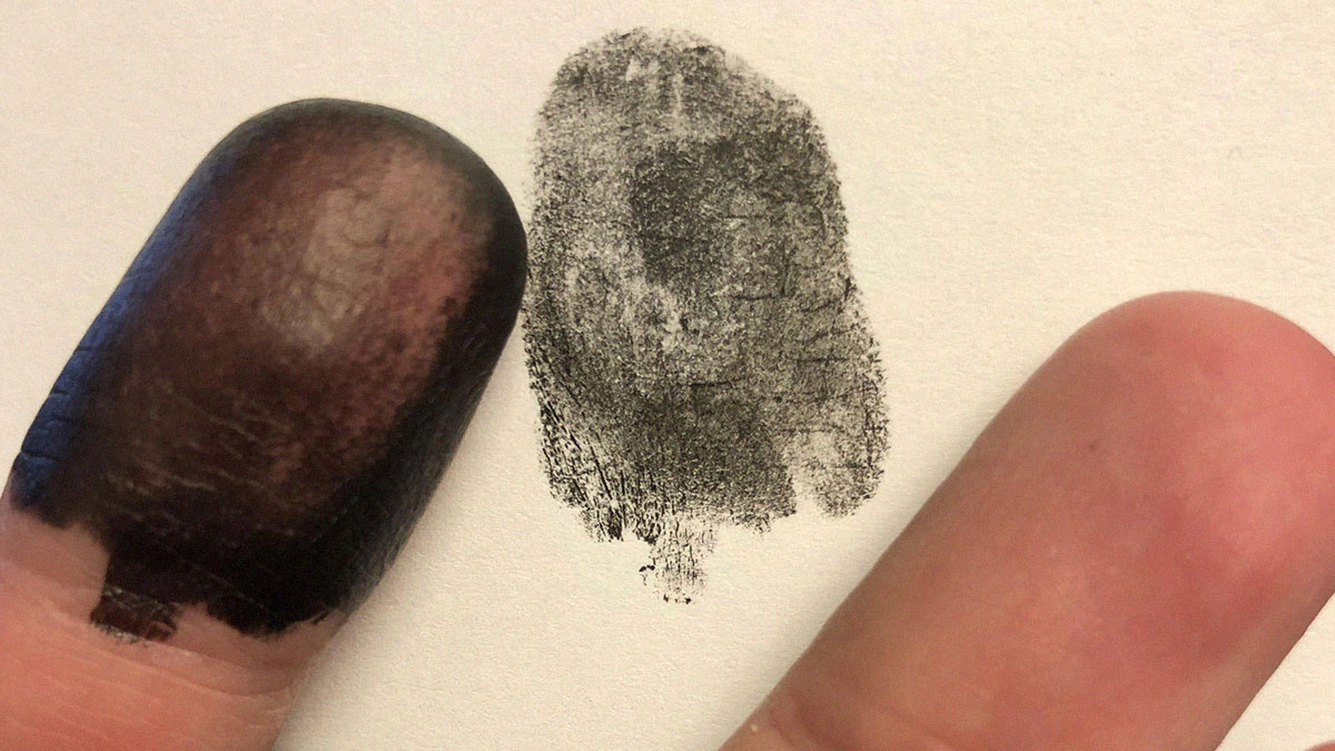 Close-up of an extra finger with a black fingerprint inked next to a normal finger on white paper.