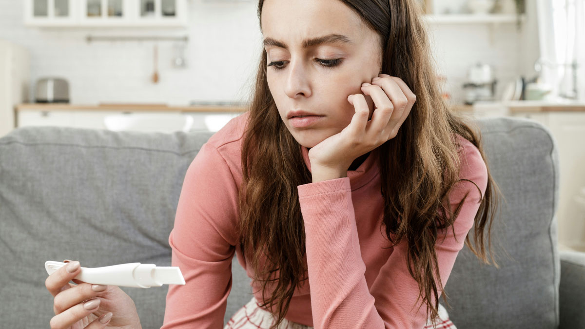 Teen girl sitting on couch looking at a pregnancy test, feeling conflicted about keeping her baby and facing lack of support from dad