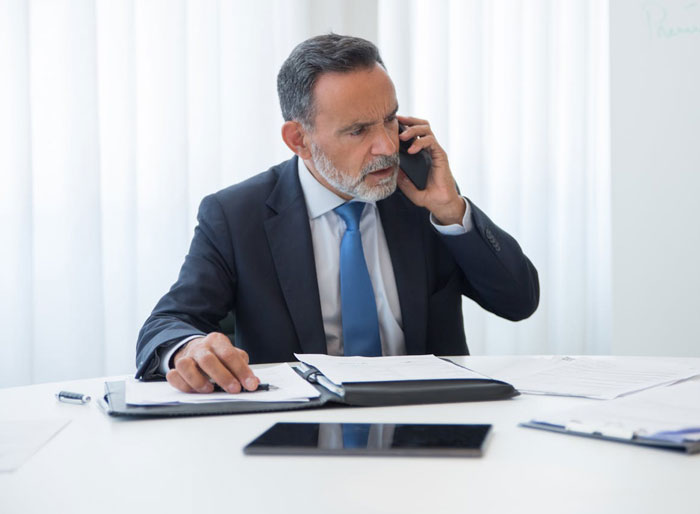 Middle-aged businessman in a suit and tie looking concerned while speaking on the phone at his office desk Middle-aged businessman in a suit and tie looking concerned while speaking on the phone at his office desk