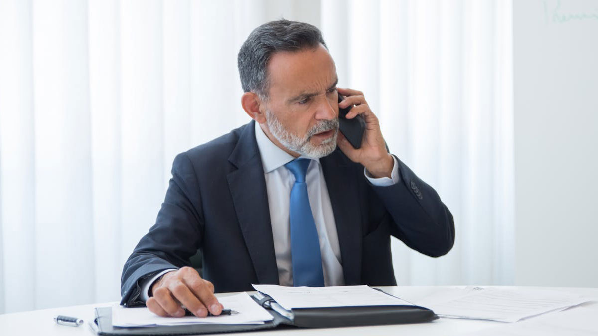 Middle-aged boss in a suit making a tense phone call at a desk, illustrating workplace conflict and difficult management.