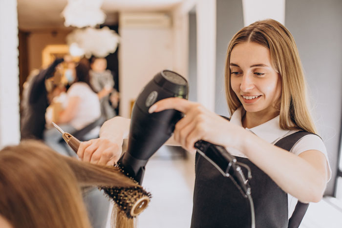 Young stylist providing free salon service marriage hairstyling with hairdryer and round brush in a bright salon setting.