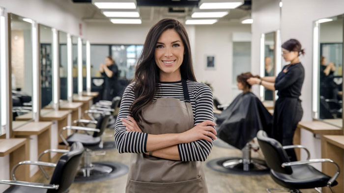 Young female stylist in a free salon service setting smiling confidently with clients getting hair treatments in the background
