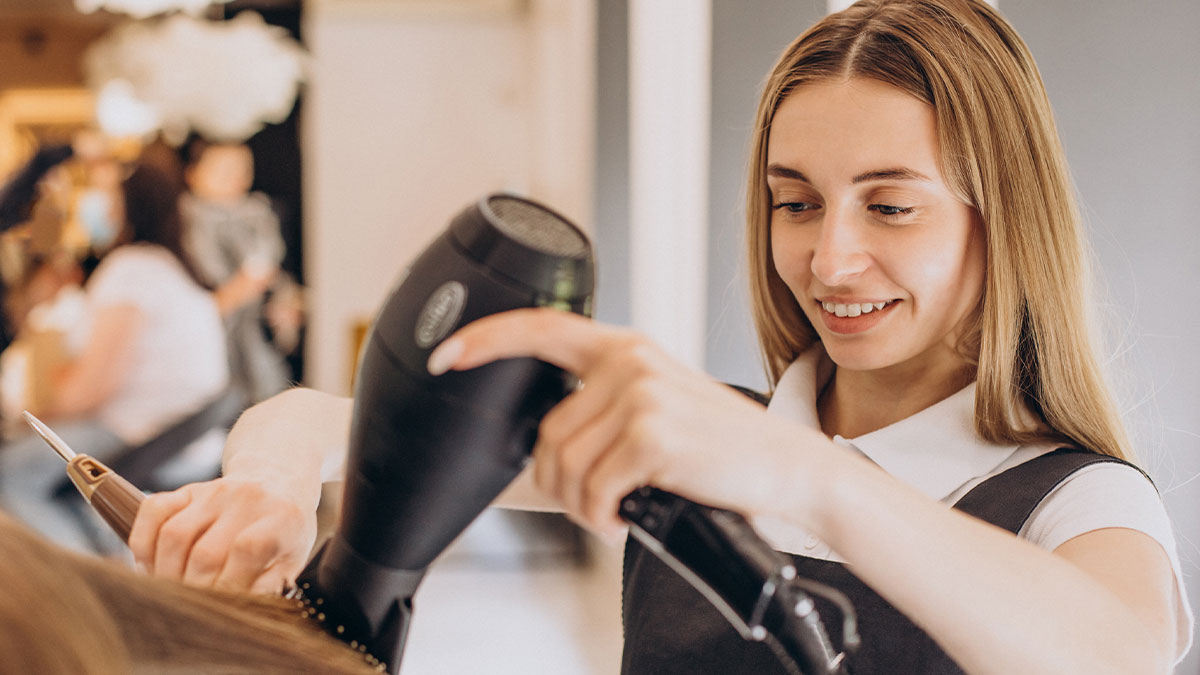 Young hairstylist blow-drying a clientu2019s hair in a salon, highlighting future DIL styling and refusing free work.