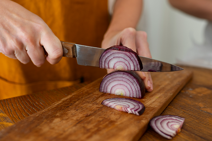 Woman slicing red onion on a wooden board with a knife, preparing ingredients in a kitchen setting. Woman slicing red onion on a wooden board with a knife, preparing ingredients in a kitchen setting.