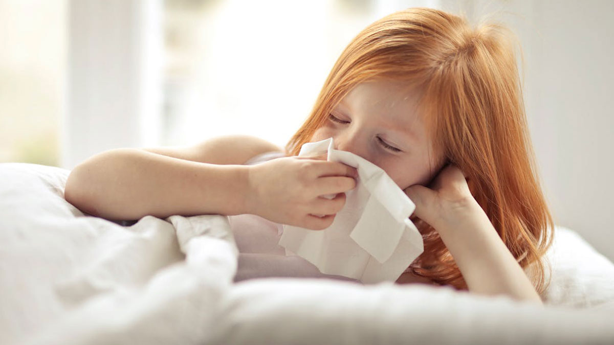 Red-haired child lying in bed blowing nose into tissue, illustrating relatable tweets making you chuckle even without kids.