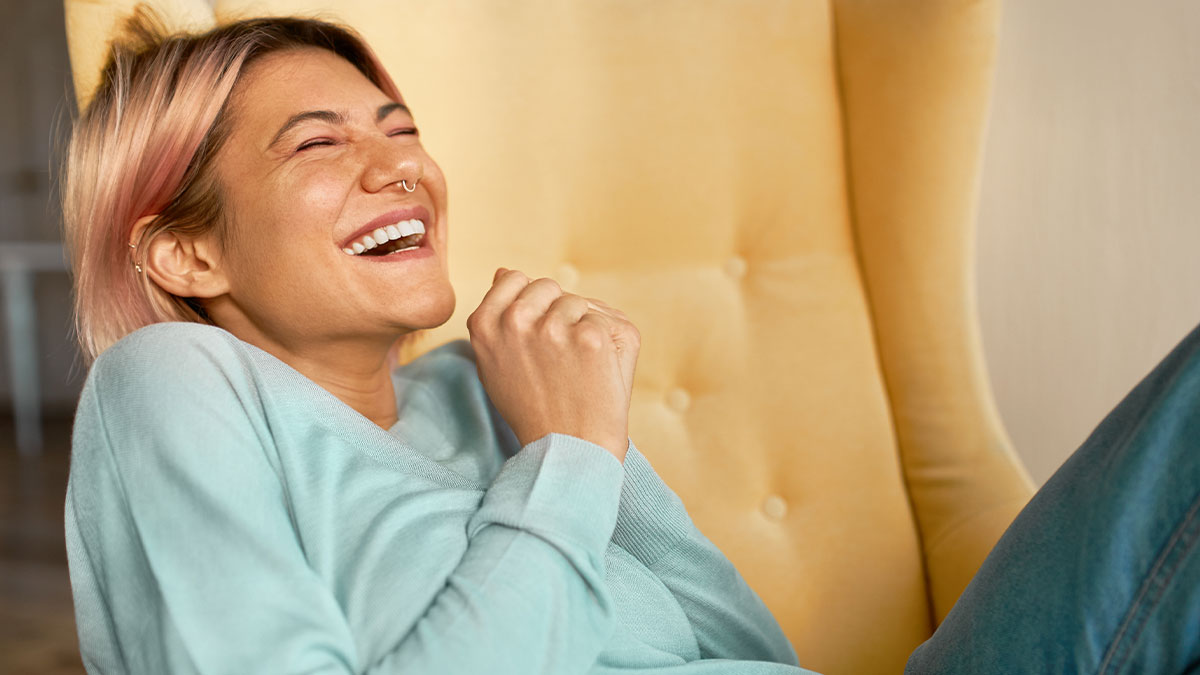 Person with pink hair laughing joyfully while relaxing on a yellow chair, enjoying random memes that bring laughter.