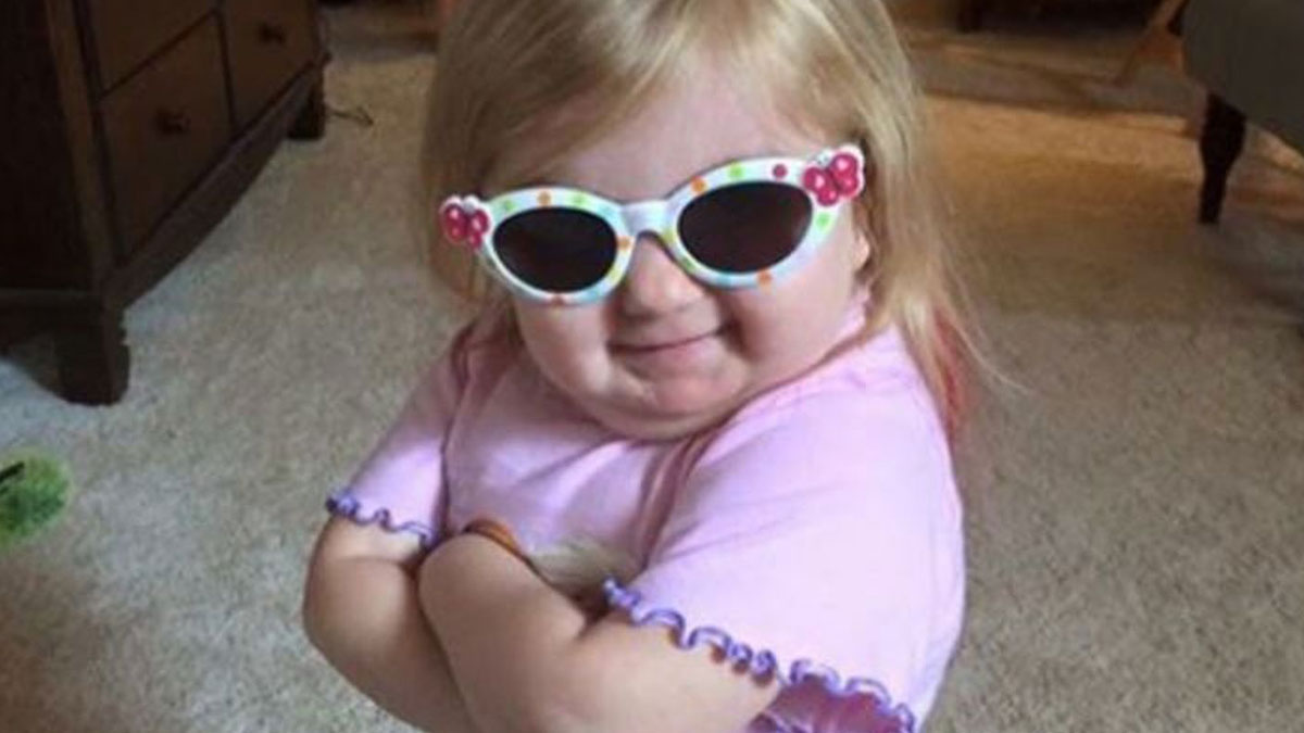 Toddler wearing colorful sunglasses and a pink shirt, smiling confidently with arms crossed, capturing cool mom parenting vibes.