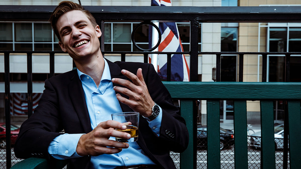 Young man dressed smartly laughing with a drink in hand outside British pub, capturing chaotic British memes vibe.