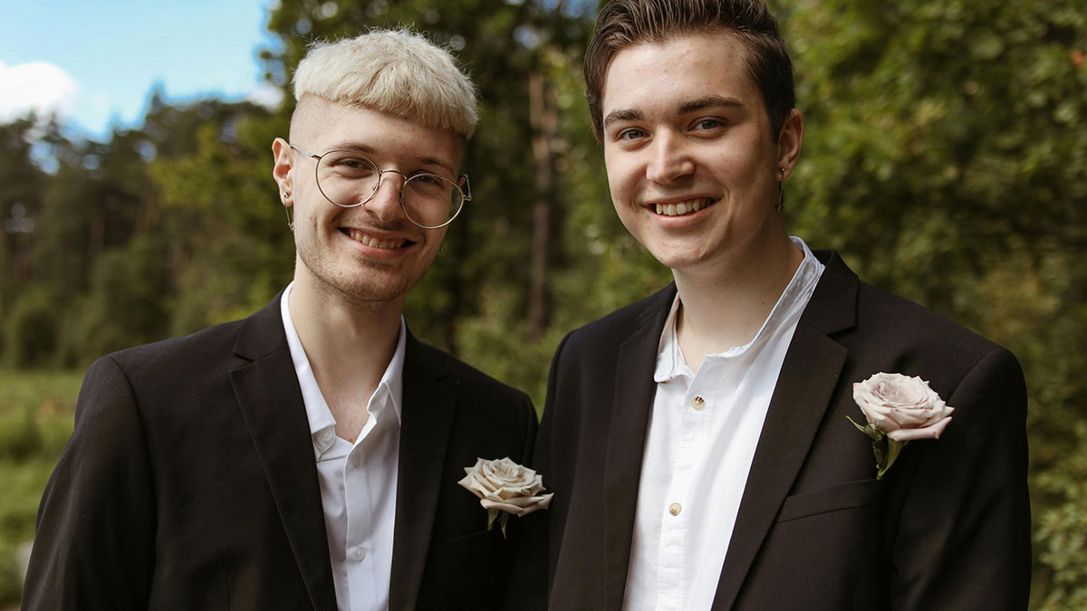 Two young men smiling outdoors at a wedding, one wearing glasses, both with boutonnieres on their black jackets.