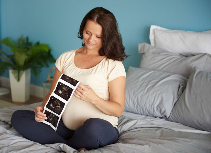 Pregnant sister sitting on bed, holding ultrasound images, reflecting on genetic condition with a calm and thoughtful expression. Pregnant sister sitting on bed, holding ultrasound images, reflecting on genetic condition with a calm and thoughtful expression.