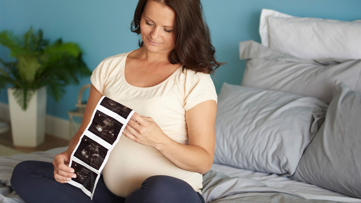 Pregnant woman sitting on bed looking at ultrasound images, reflecting on genetic disease challenges.
