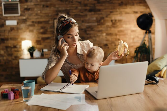 Woman multitasking with a baby, phone, laptop, and notebook highlighting gender-based double standards in work and parenting.
