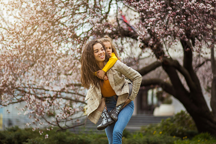 Smiling woman giving piggyback ride to child near blooming trees, capturing a joyful family moment outside. Smiling woman giving piggyback ride to child near blooming trees, capturing a joyful family moment outside.