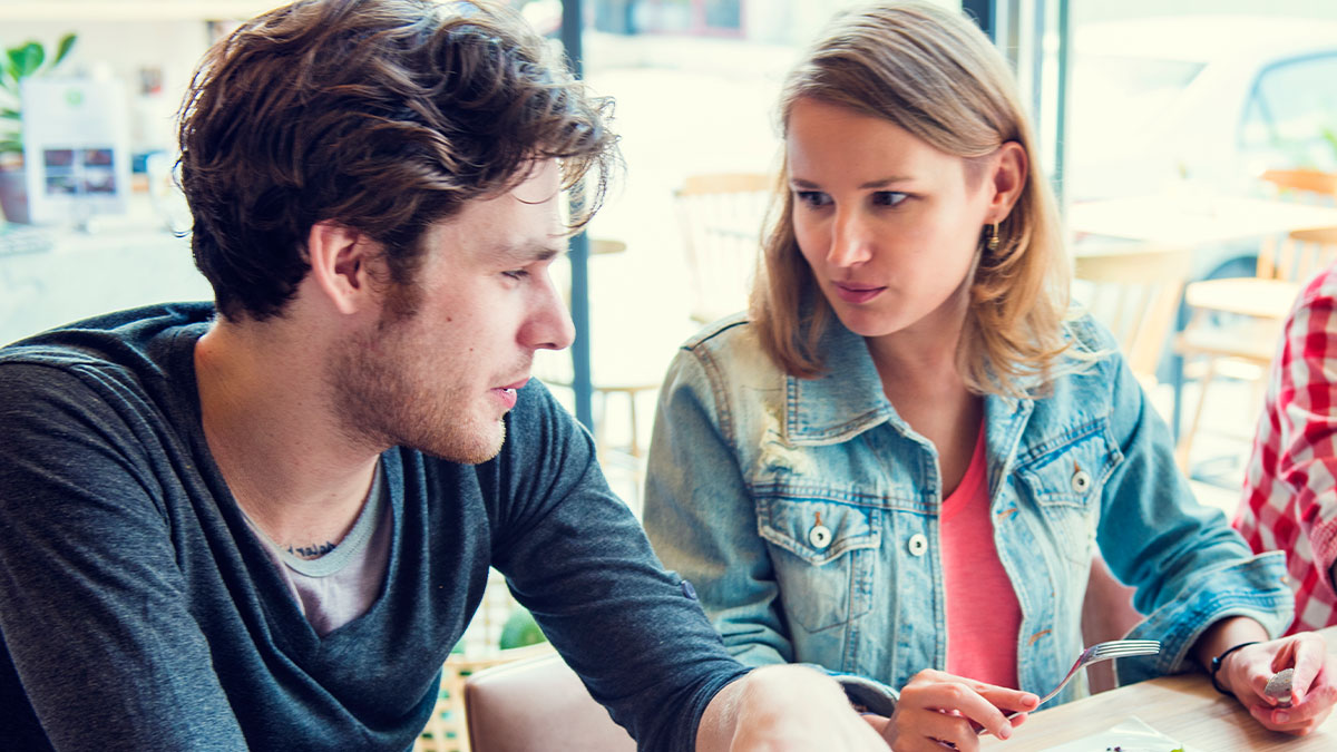 Man and upset woman sitting at a table, woman holding fork with peanut butter cake, man unable to eat it at work.