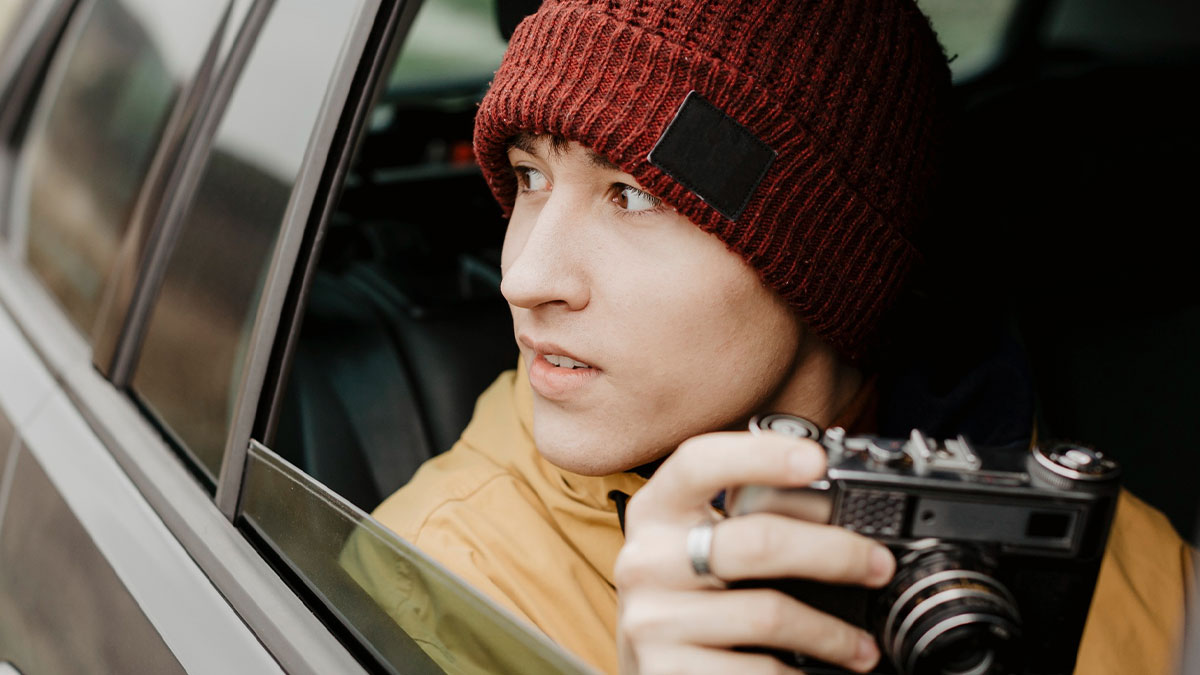Teen holding vintage camera looking out car window, capturing moments during portable darkroom adventure at surplus auction.
