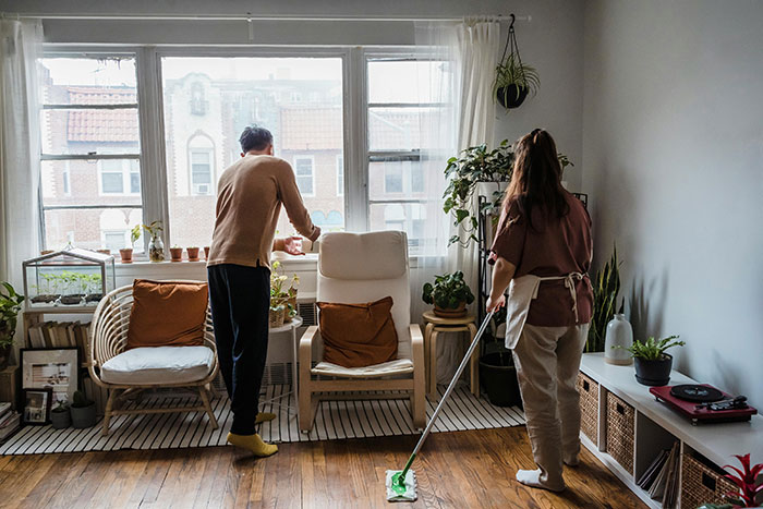 Woman mopping floor as man looks outside window in cozy living room, reflecting grandma threaten CPS unborn baby tension. Woman mopping floor as man looks outside window in cozy living room, reflecting grandma threaten CPS unborn baby tension.