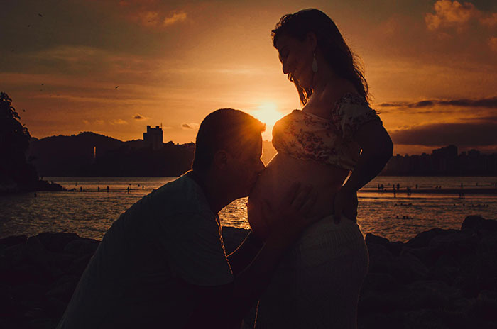 Couple at sunset with man kissing pregnant woman’s belly symbolizing unborn baby in a family setting near water. Couple at sunset with man kissing pregnant woman’s belly symbolizing unborn baby in a family setting near water.