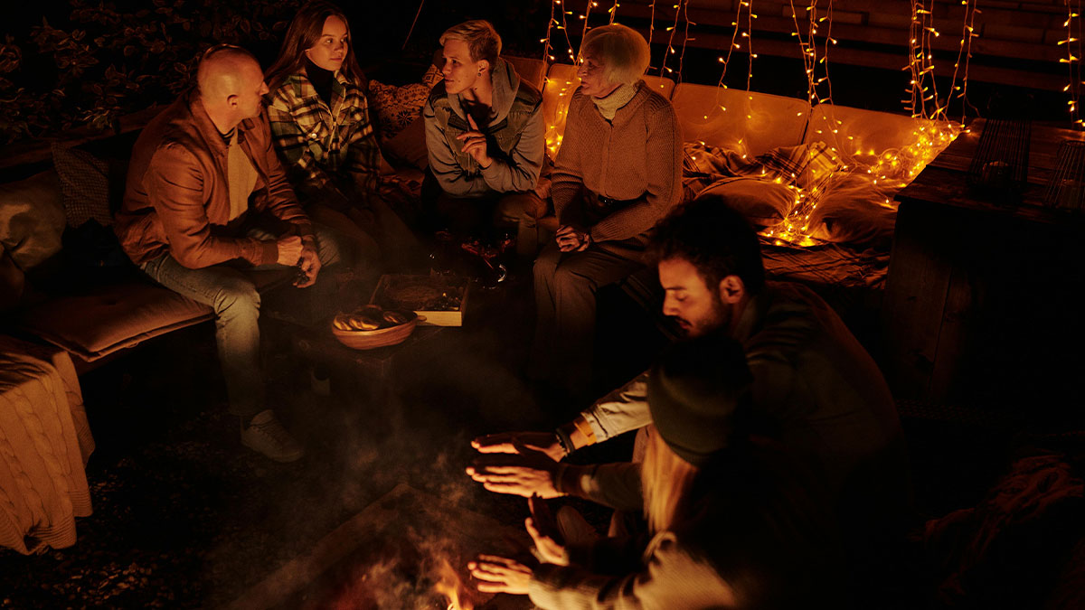Group of people including a grandma gathered around a fire outdoors, warm lighting creating an intimate atmosphere.