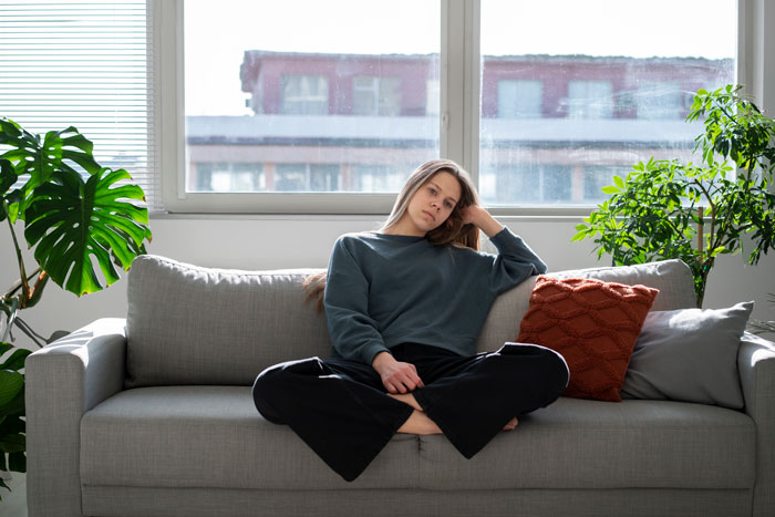 Young woman sitting on a gray sofa looking pensive, representing reluctance as future caregiver for autistic stepbrother. Young woman sitting on a gray sofa looking pensive, representing reluctance as future caregiver for autistic stepbrother.