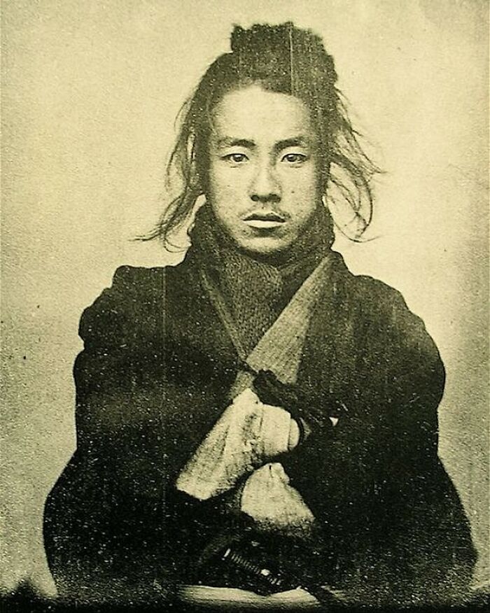 Victorian man with long hair wearing traditional clothing, captured in a vintage black and white portrait.