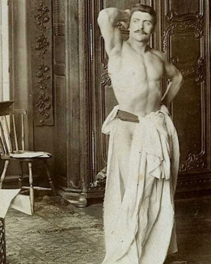 Victorian handsome man posing shirtless with a draped cloth in a vintage room with wooden furniture and ornate decor.