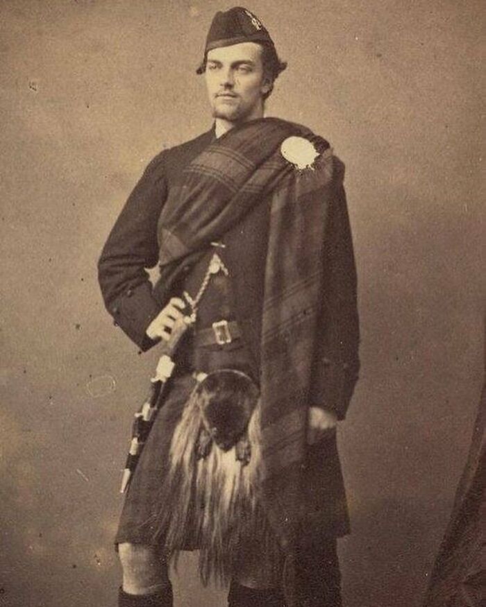 Young Victorian man dressed in traditional Scottish attire, standing confidently with hand on hip in a vintage sepia photo.