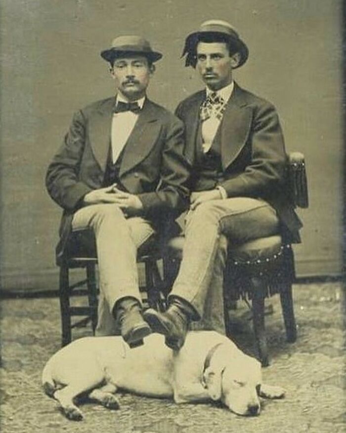 Two handsome Victorian men seated on chairs, dressed in period attire with hats, accompanied by a dog lying on the floor.