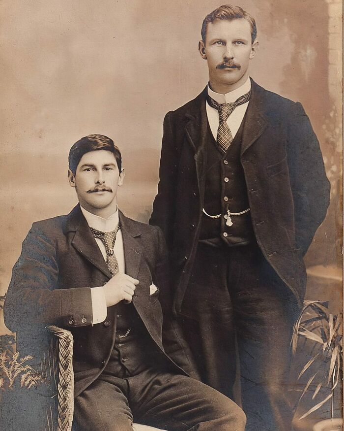 Two handsome Victorian men with mustaches wearing formal three-piece suits and ties in a vintage sepia portrait.