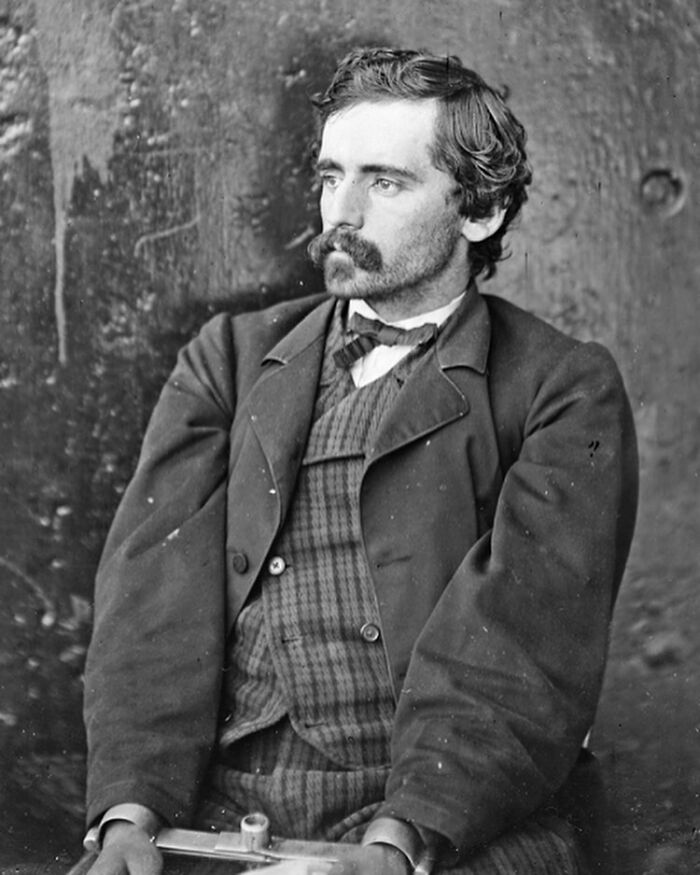 Victorian man with mustache and bow tie wearing a suit, sitting and looking thoughtfully to the side in a vintage black and white photo.