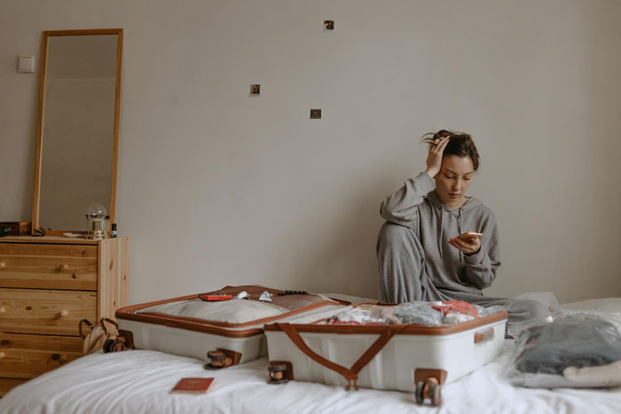 Childfree woman sitting on bed, frustrated and looking at phone, next to open suitcase while packing for vacation. Childfree woman sitting on bed, frustrated and looking at phone, next to open suitcase while packing for vacation.