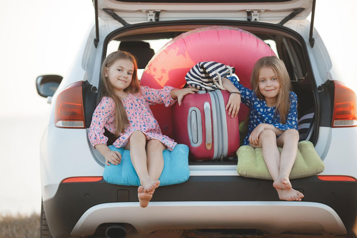 Two young girls sitting on cushions in an open car trunk filled with travel luggage on vacation trip. Two young girls sitting on cushions in an open car trunk filled with travel luggage on vacation trip.