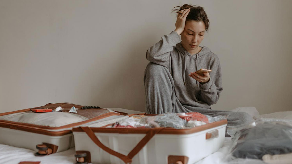 Childfree woman in gray loungewear sitting on bed, checking phone next to open suitcase, reflecting on vacation escape.