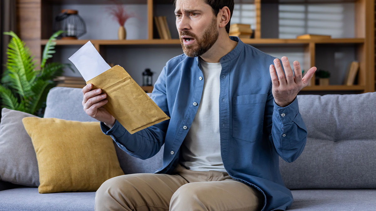 Man sitting on couch looking frustrated while holding an envelope, dealing with petty HOA follow ups.