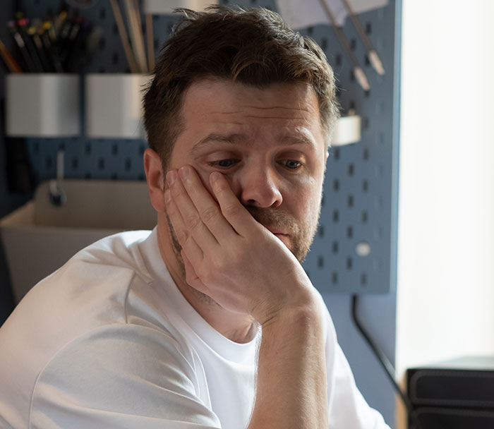 Man in a white shirt touching his face, showing a body quirk in a thoughtful pose indoors with soft lighting.