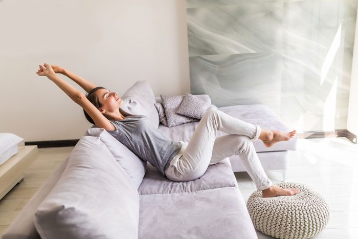 Young woman relaxing on a modern couch, stretching comfortably in a bright, minimalistic living room setting.