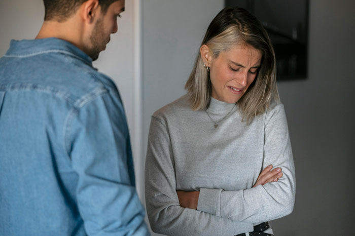 Woman in gray sweater crying with arms crossed as husband in denim jacket looks on, highlighting hubby mocks wife over mispronunciation. Woman in gray sweater crying with arms crossed as husband in denim jacket looks on, highlighting hubby mocks wife over mispronunciation.