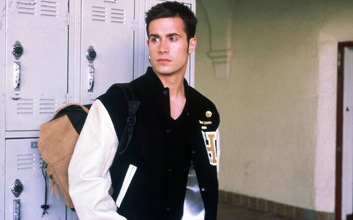 Young male actor posing by school lockers wearing a letterman jacket, embodying classic heartthrob style.