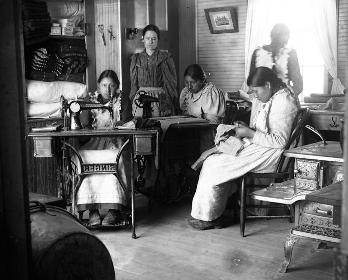 Native American girls in the 1910s at a boarding school sewing and engaged in handiwork inside a classroom.