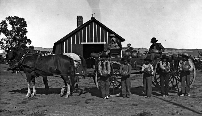 Children at a Native American boarding school in the 1910s standing with pumpkins by a horse-drawn wagon in front of a barn.